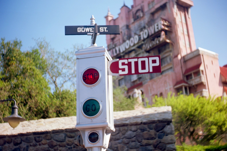 A vintage-style traffic signal at the intersection of Gower Street, displaying a prominent “STOP” sign with illuminated red and green lights. The Hollywood Tower Hotel, famously known as the Tower of Terror, looms ominously in the background at Disney's Hollywood Studios. The iconic attraction's weathered architecture and eerie ambiance are contrasted by the bright blue sky and lush greenery surrounding the scene. This photograph was captured on an Argus C3 camera using Kodak Ektar film, adding a nostalgic filmic quality. Captured on an Argus C3 camera with Kodak Ektar film. A Work by John Paglia.