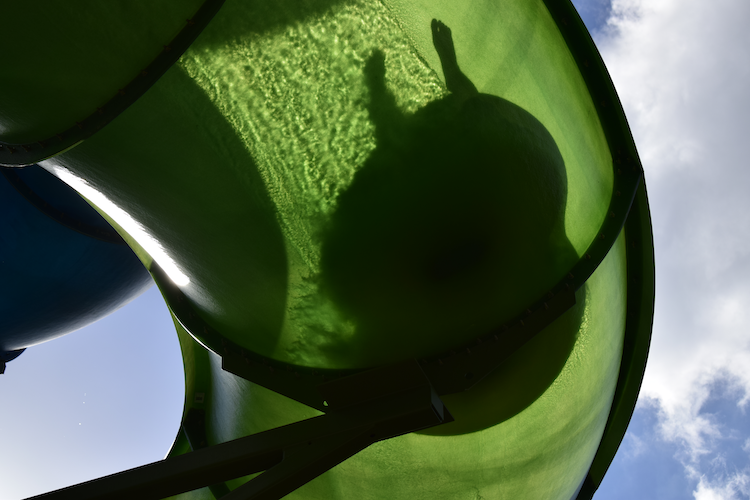 A unique perspective of the Tahniwah Tubes water slide at Universal's Volcano Bay, captured from below. The translucent green slide glows under the sunlight, revealing the silhouette of a rider as they zoom through the tube. The vibrant green contrasts against the bright blue sky with scattered clouds in the background, emphasizing the thrilling and immersive water park experience. Captured on a Nikon D5300 digital camera. A Work by John Paglia.