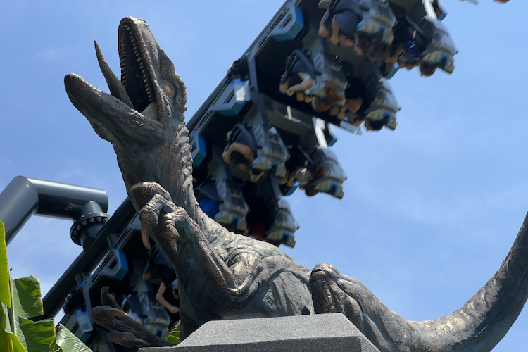 A dynamic view of a Velociraptor statue at Universal's Islands of Adventure, standing as a menacing centerpiece for the Jurassic World VelociCoaster. In the background, the coaster train soars through the sky, filled with riders enjoying the thrilling inverted track. The intricate detail of the raptor's claws and teeth contrasts with the sleek, high-tech design of the coaster vehicles. Surrounded by tropical foliage, this photograph captures the immersive blend of adventure and excitement in the Jurassic World-themed area. Shot on iPhone. A Work by John Paglia.