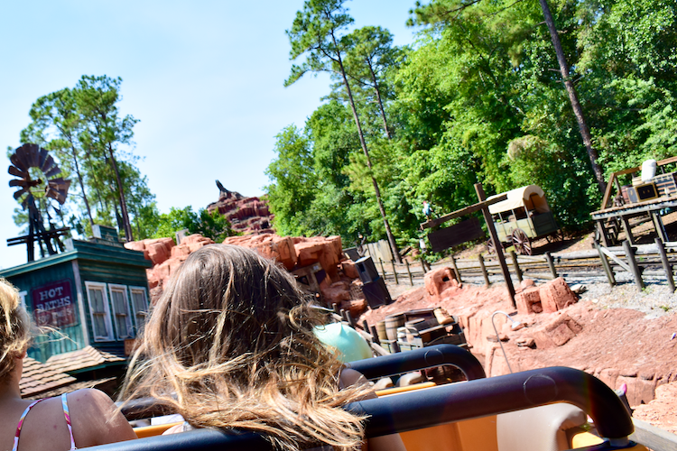 A vibrant photograph taken while riding Big Thunder Mountain Railroad at Magic Kingdom in Walt Disney World, Orlando, Florida. The image captures the dynamic movement of the roller coaster with the iconic rustic landscape, featuring red rock formations, a green forested backdrop, and a windmill. In the distance, Splash Mountain can be seen, adding depth to the Disney theme park experience. The back of two riders with flowing hair emphasizes the thrilling nature of the ride. Captured on a Nikon D5300 digital camera. A Work by John Paglia.