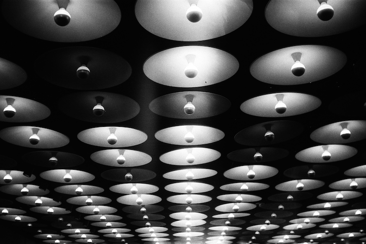 Black-and-white photograph of the iconic lobby ceiling at 945 Madison Avenue, The Breuer Building, showcasing an array of circular light fixtures. Each fixture features a central light bulb suspended in the middle of a concave reflective dish, creating a rhythmic pattern of light and shadow across the space. The repetitive geometric design emphasizes modernist architectural elements and the innovative design ethos of Marcel Breuer. The image captures the interplay of light and texture, highlighting the architectural significance of the building, originally constructed as the Whitney Museum of American Art and now housing the Frick Madison. Captured on an Argus C3 camera with Ilford HP5 Plus film. A work by John Paglia.