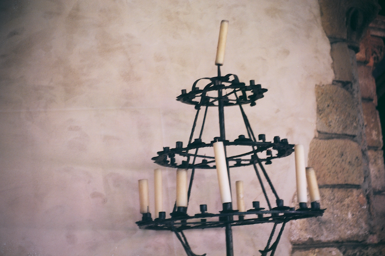 A wrought iron candelabra located at The Met Cloisters, featuring multiple tiers of circular holders for candles. The structure holds several tall, unlit candles, evoking a medieval aesthetic. The background consists of a textured stone wall with warm hues, complementing the rustic charm of the candelabra. The composition highlights the interplay of light and shadow, creating a timeless and serene atmosphere reflective of the historical setting. Captured on an Argus C3 camera with Kodak Ektar film. A work by John Paglia.