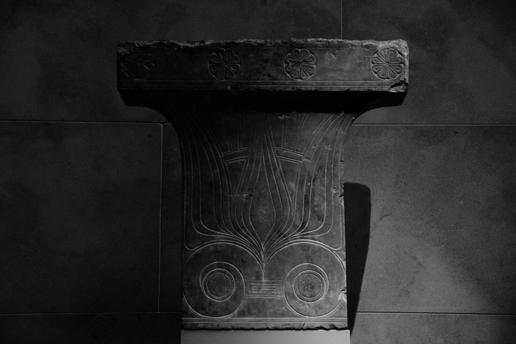 A carved Ionic capital from the ancient Greek period, showcasing intricate floral and geometric motifs. The capital features a central palmette design and volutes, symbolizing classical architectural refinement. The dark stone is illuminated dramatically, emphasizing the delicate carving details and the weathered texture of the artifact. This piece, housed at The Metropolitan Museum of Art, exemplifies the elegance and craftsmanship of ancient Greek artistry. Captured on a Nikon D5300 digital camera. A work by John Paglia.