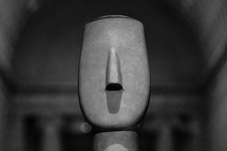 A close-up photograph of a Cycladic sculpture at The Metropolitan Museum of Art. The marble figure showcases minimalist features, including a stylized face with a long, narrow nose and an open mouth set against a smooth, elongated head. The lighting emphasizes the polished texture and subtle curves of the ancient artifact, which reflects the iconic simplicity and elegance of Cycladic art. The blurred background adds depth to the image, focusing attention on the timeless craftsmanship of the piece. Captured on a Nikon D5300 digital camera. A work by John Paglia.