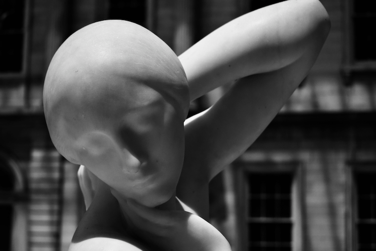 The marble sculpture Fragilina by Attilio Piccirilli, capturing a delicate and introspective pose of a female figure with her head tilted and hand gently resting on her neck. The smooth, polished surface of the sculpture reflects the artist's mastery of form and texture. The background, blurred yet showing architectural details, contrasts with the soft contours of the sculpture, emphasizing its serene beauty and timeless elegance. Displayed in natural light, the play of shadows highlights the intricate details of this exquisite work of art. Captured on a Nikon D5300 digital camera. A work by John Paglia.