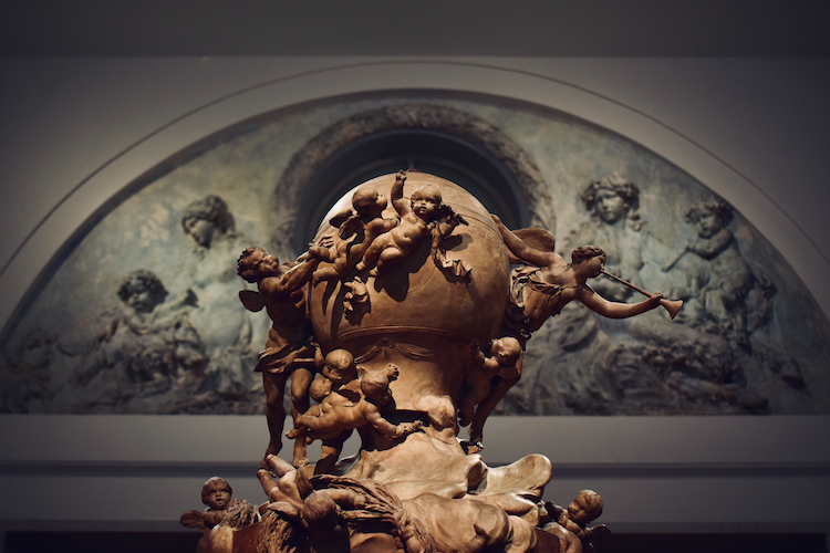 An intricate Italian Baroque sculpture displayed in Gallery 551 at The Metropolitan Museum of Art in New York City. The centerpiece features cherubic figures dynamically arranged around a spherical globe, exuding movement and dramatic expression. The background showcases an arched fresco with ethereal figures, complementing the sculpture's classical Baroque artistry. The warm tones and intricate details highlight the ornate craftsmanship characteristic of the Italian Baroque period. Captured on a Nikon D5300 digital camera. A work by John Paglia.