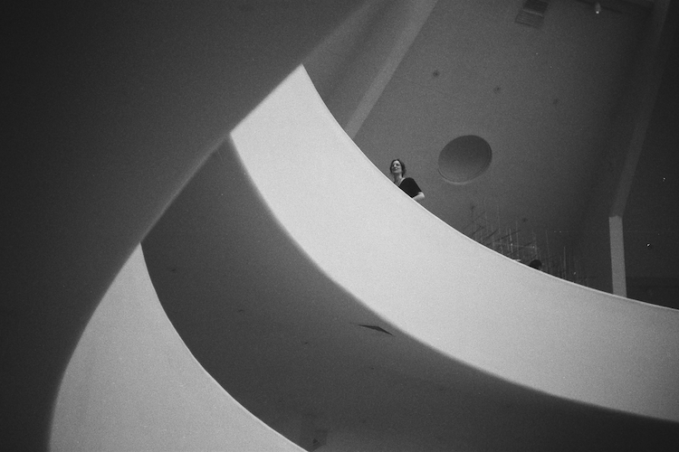 Black and white photograph of the Guggenheim Museum's iconic architecture, featuring a woman standing on a curved balcony. The sweeping lines, geometric shapes, and dramatic contrast of light and shadow highlight the museum's modern design. Capturing a moment of stillness within the architectural masterpiece, this image reflects the beauty of minimalist spaces and thoughtful composition. Perfect for showcasing fine art photography, museum interiors, and architectural design. Captured on an Argus C3 camera with Ilford HP5 Plus film. A work by John Paglia.