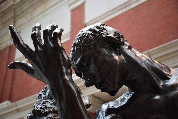 A close-up of a bronze statue from Auguste Rodin's “The Burghers of Calais,” located at The Metropolitan Museum of Art. The image captures an expressive figure in emotional turmoil, with its head bowed and a hand raised dramatically. The detailed texture of the sculpture highlights the raw emotion and vulnerability of the subject, emphasizing the tension in the muscles and the intricacies of the face and hand. The backdrop features the museum's interior with a red brick wall, complementing the dark patina of the bronze. Captured on a Nikon D5300 digital camera. A work by John Paglia.