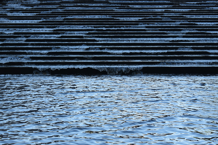 A cascading water feature at the Fondation Louis Vuitton in Paris, France. The image captures the dynamic movement of water flowing over evenly spaced horizontal steps, creating ripples and textures on the surface. The lower pool reflects the light and surrounding environment, adding a sense of depth and tranquility. This architectural detail highlights the modern and artistic design of the Fondation Louis Vuitton, seamlessly blending natural elements with contemporary structures. Captured on a Nikon D5300 digital camera. A work by John Paglia.